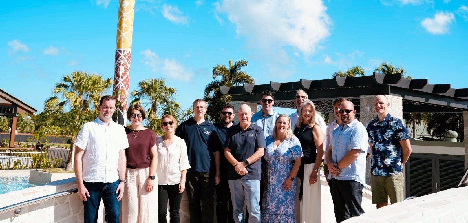 Group of people standing outdoors posing for a photo with a clear blue sky and palm trees in the background.