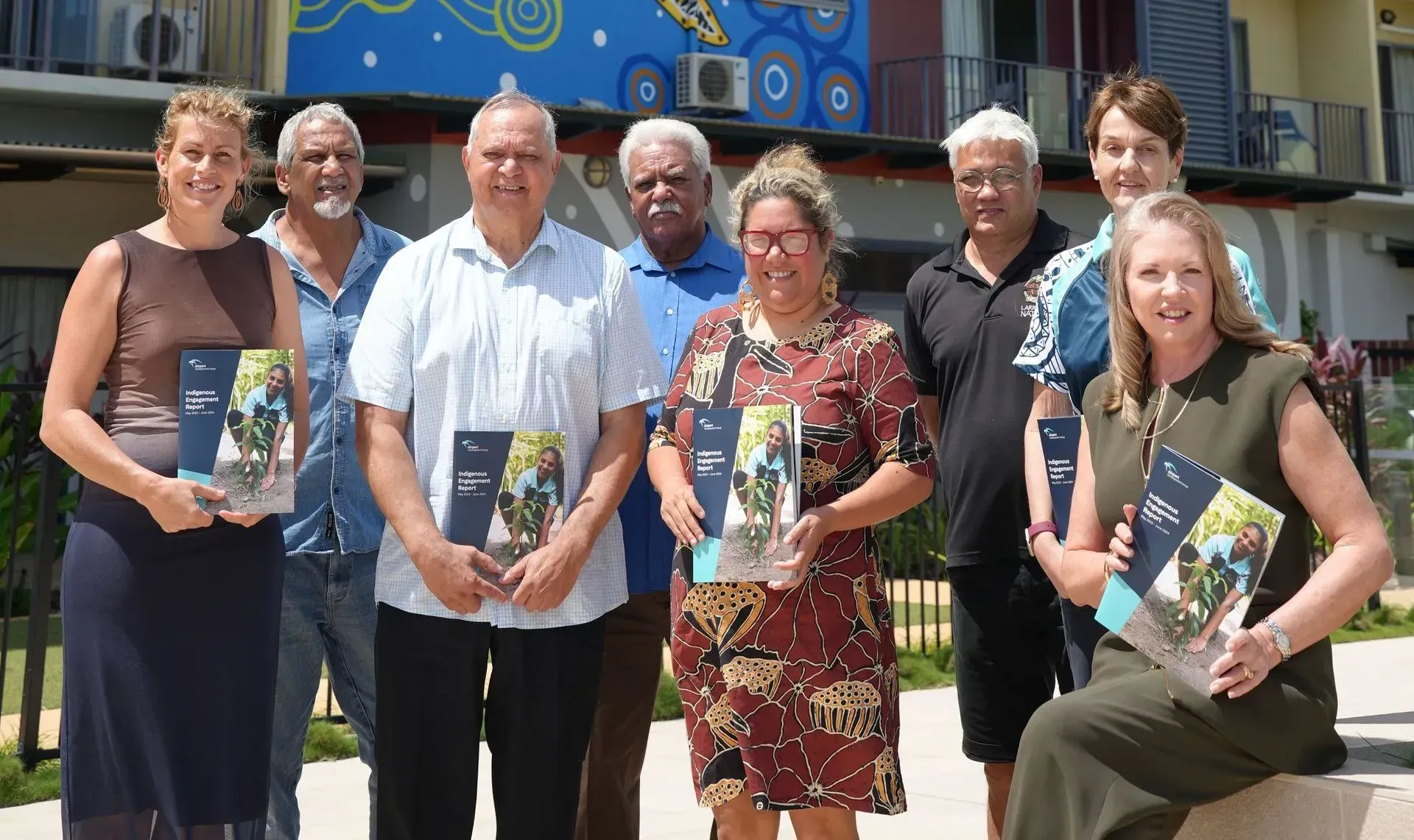 Group of people holding books outdoors, smiling, against a colorful mural.