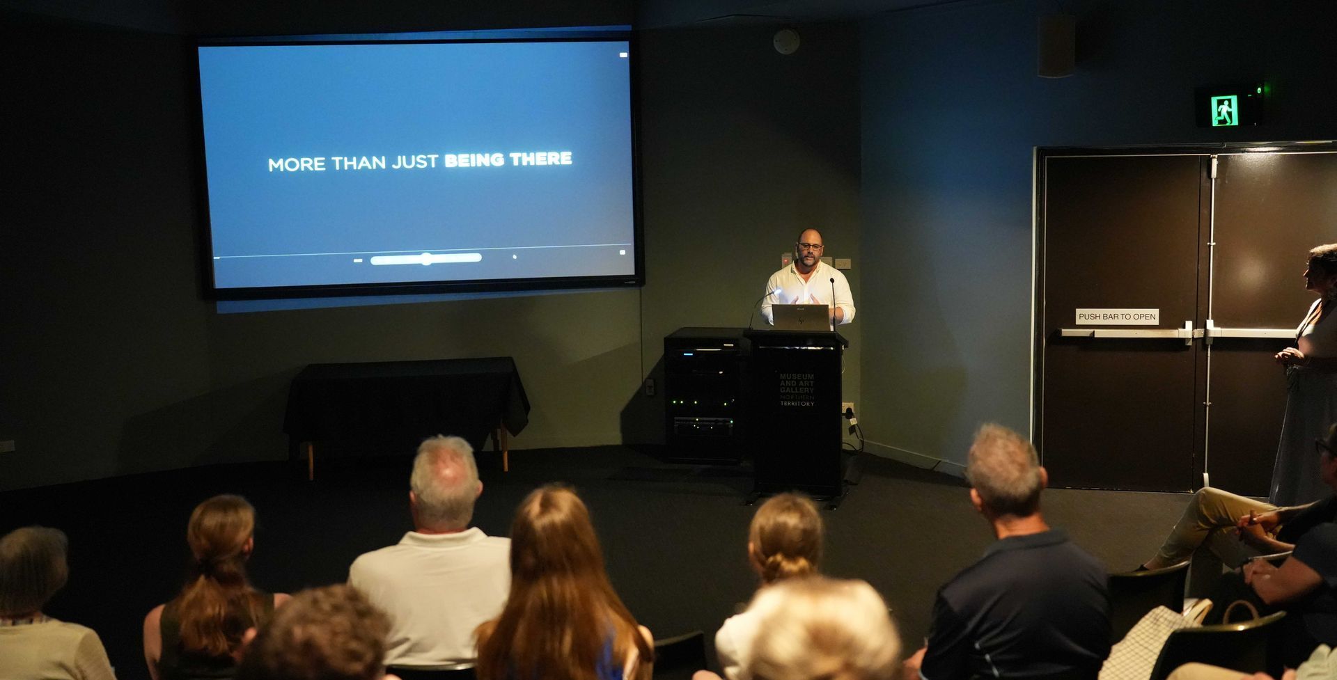 Man presenting to an audience; screen shows 