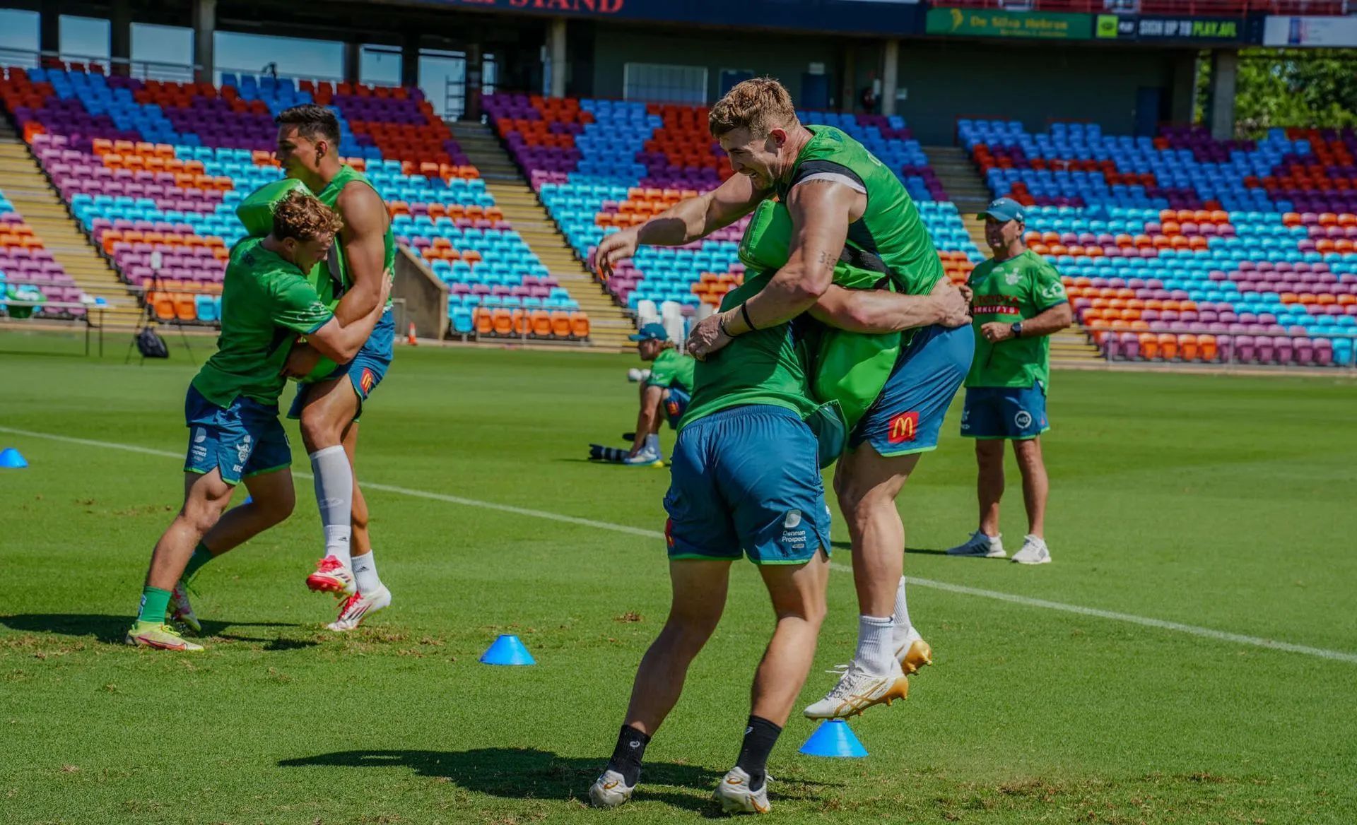 Cricket players in green and blue gear embrace on a sunny field, with stadium seating in the background.