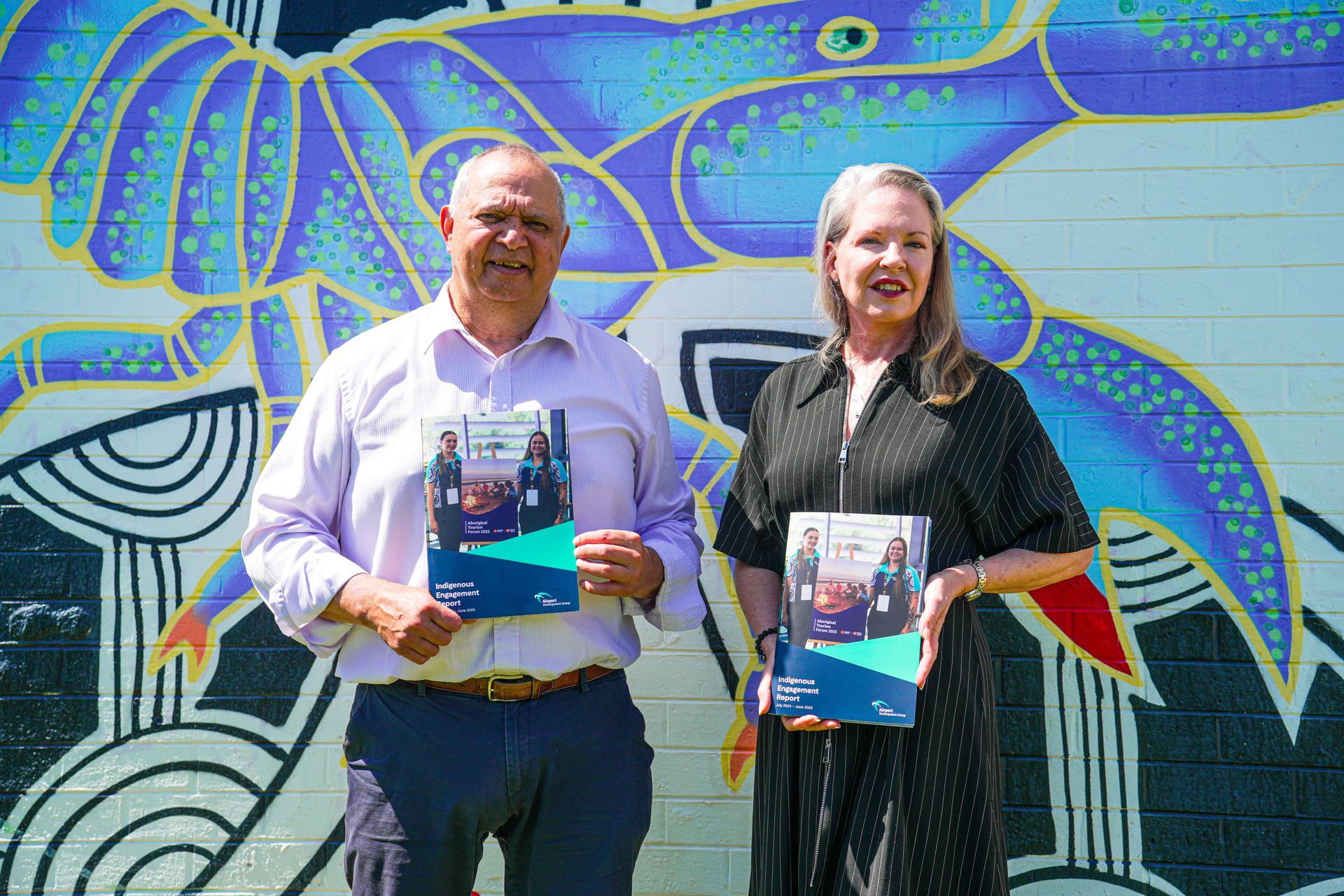 Two people holding brochures in front of a colorful mural.
