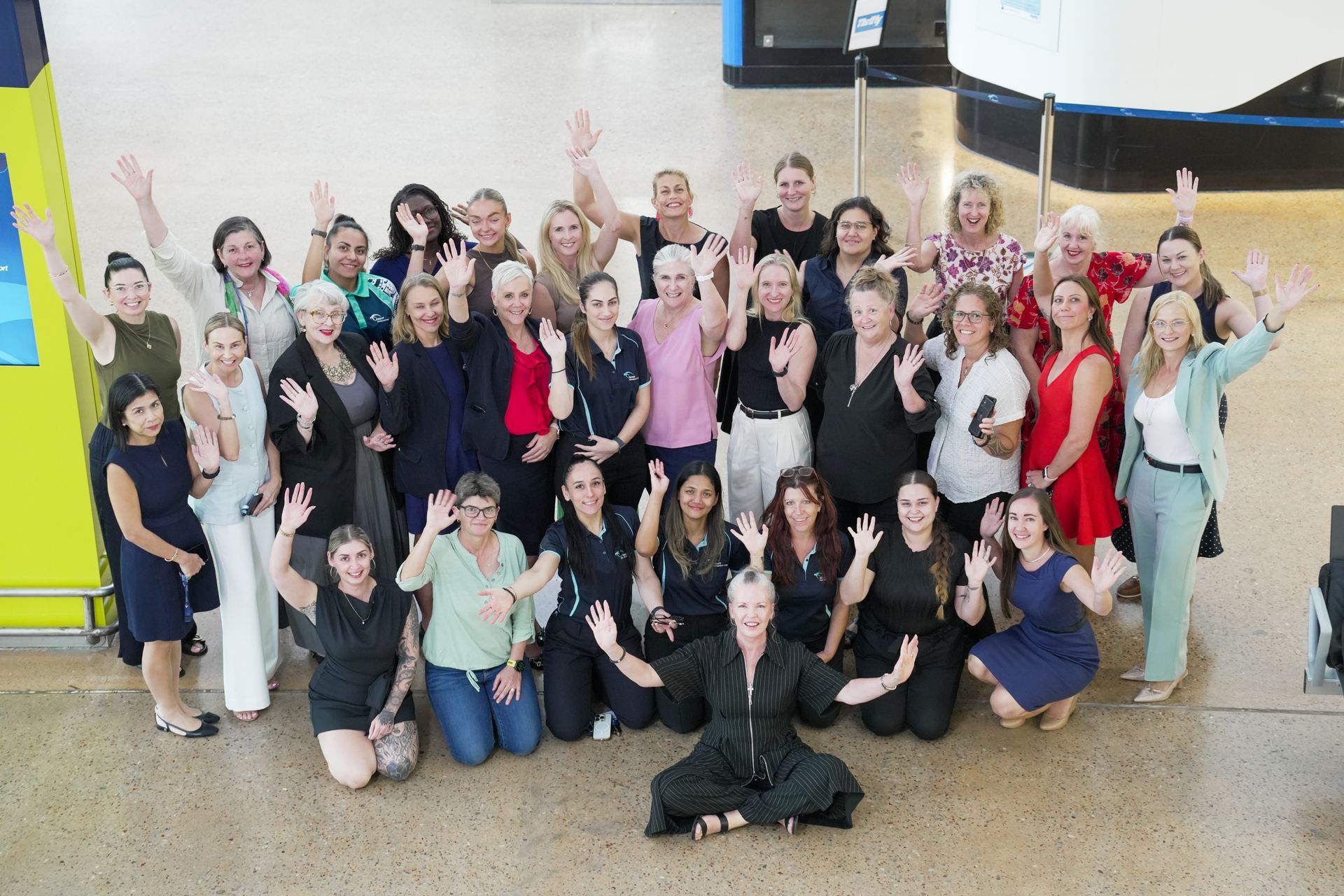 Group of women waving, posing in a building. Some are kneeling, others standing, wearing diverse clothing.