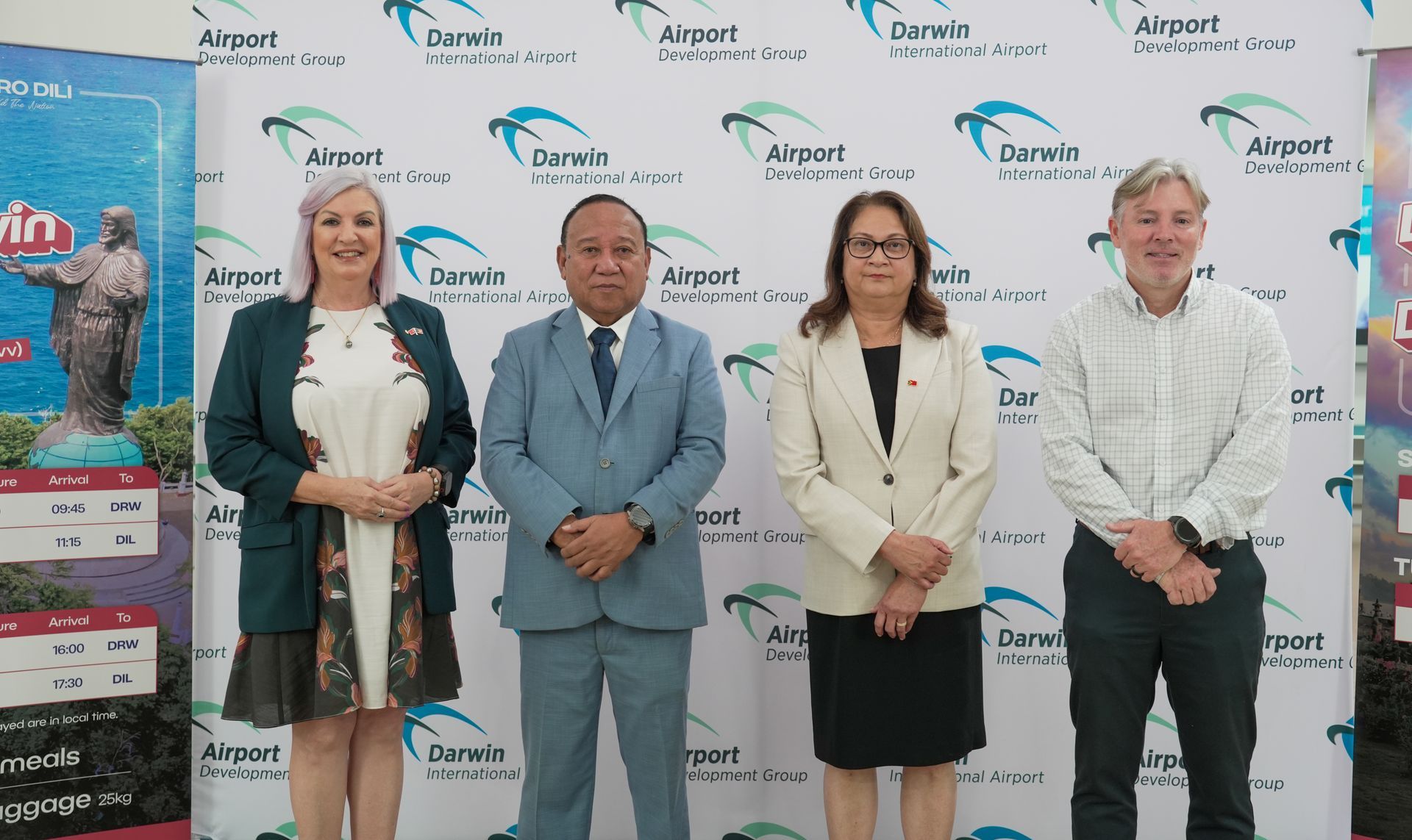 Four people stand in front of a backdrop with logos. They are smiling, wearing business attire, and arms are crossed or at their sides.