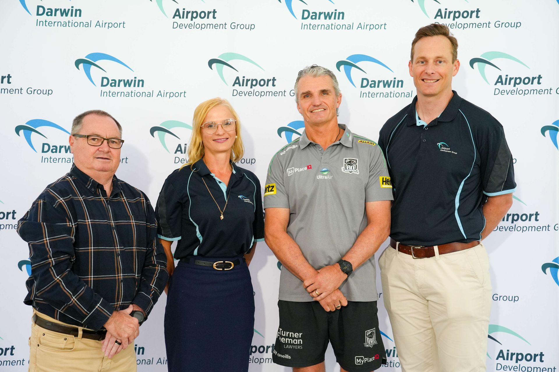 Four people stand side-by-side in front of a white backdrop with repeated Darwin International Airport logos.