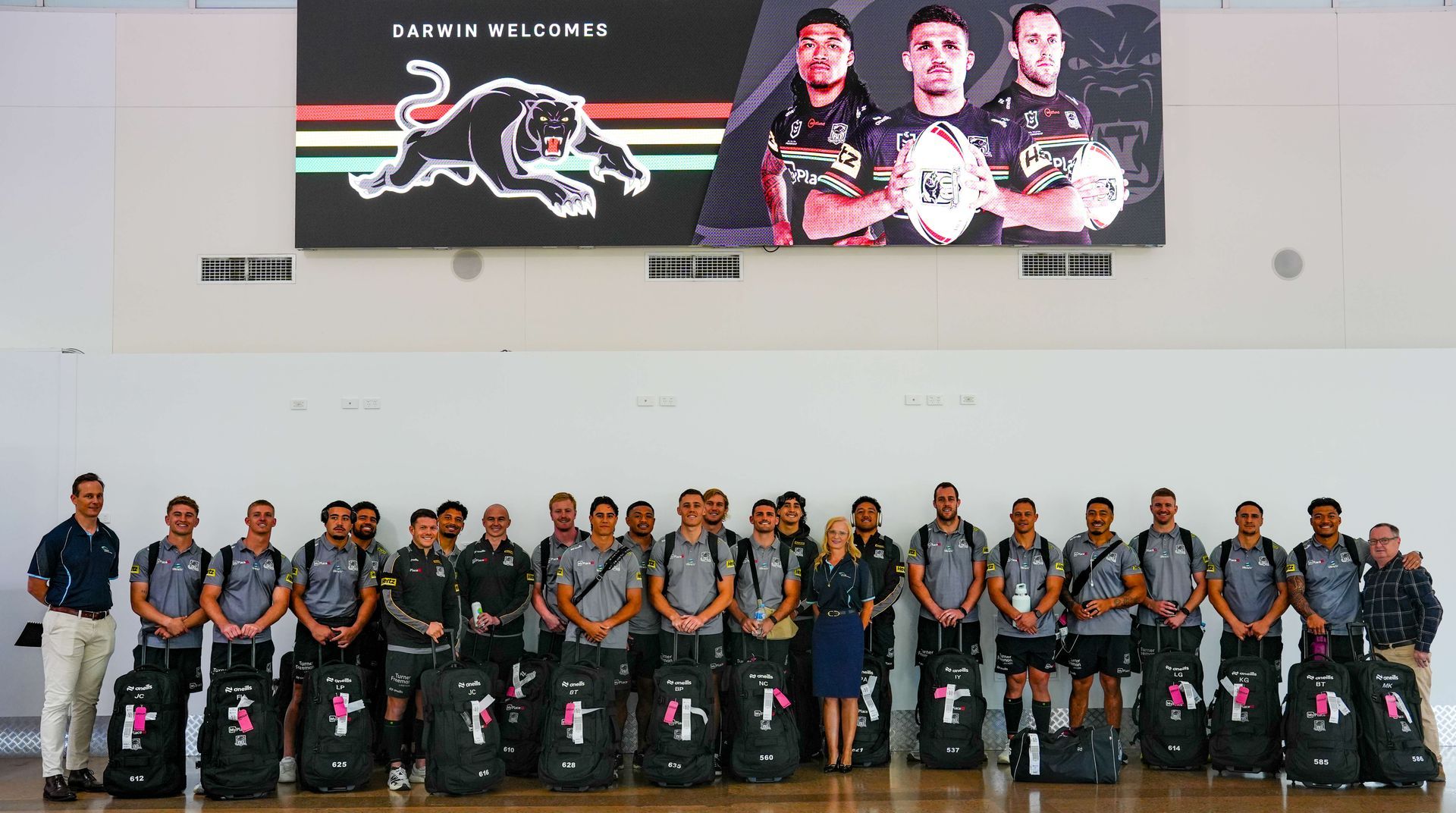 A sports team stands in a row with their travel gear before a large screen displaying their logo and three players.