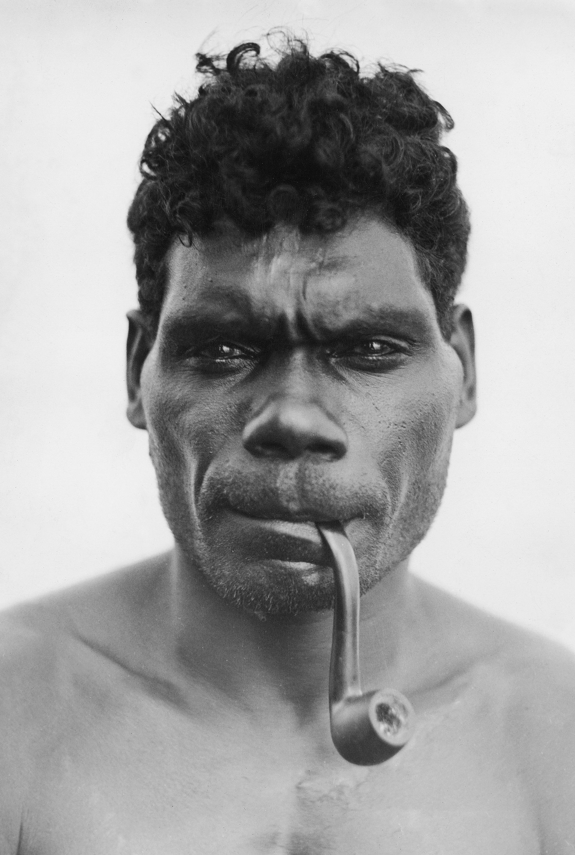 Black and white portrait of Indigenous Australian man with pipe in mouth, intense gaze.
