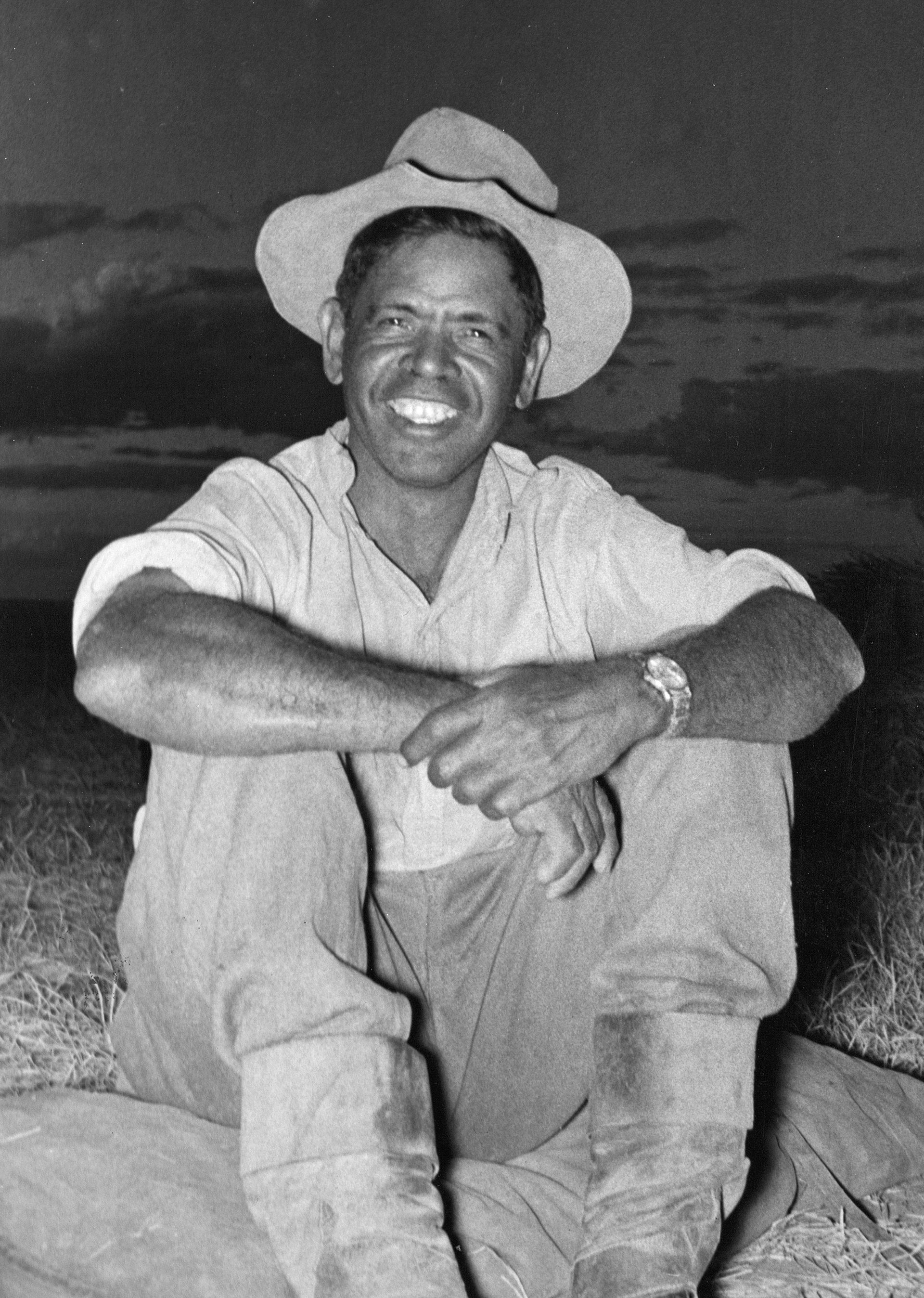 Smiling Black man in a hat, sitting outdoors, wearing work clothes, arms crossed, wristwatch visible.