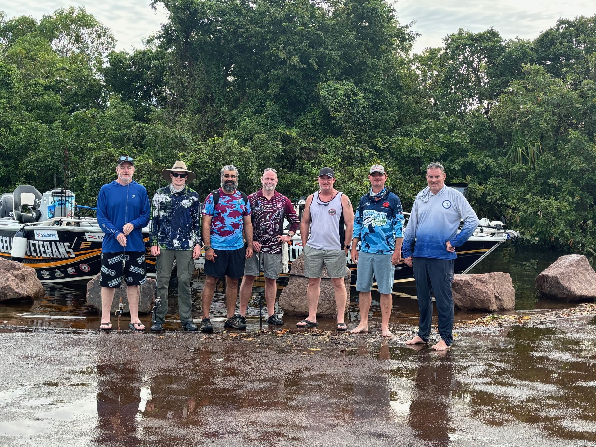 Seven people standing on a wet riverbank with kayaks, posing in rain gear and outdoor clothing
