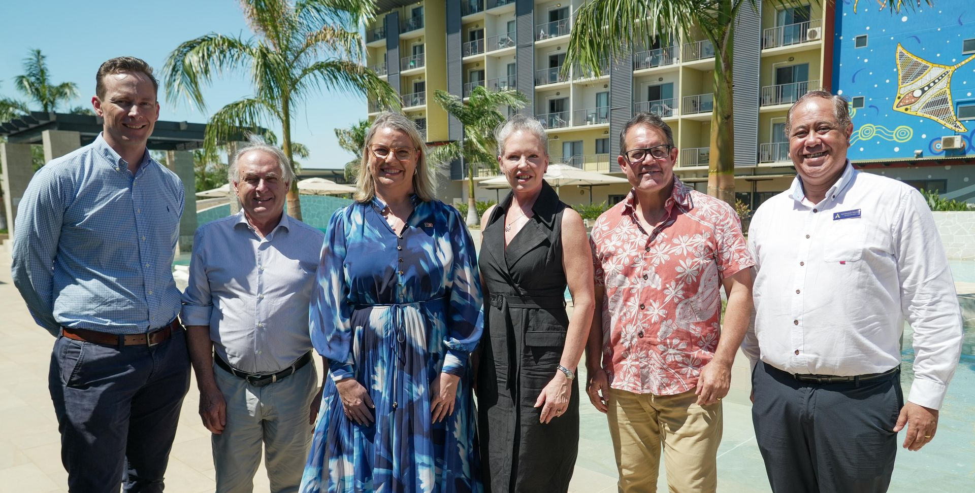 Six adults standing outdoors near a resort with palm trees and a pool.
