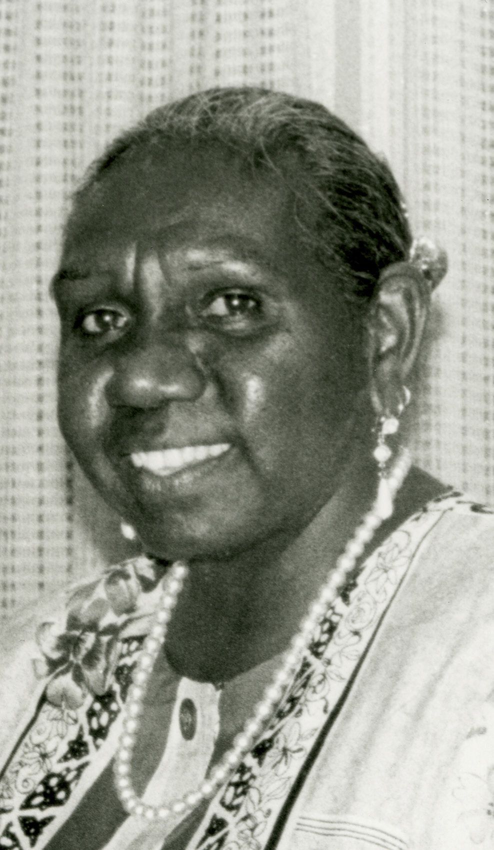 Smiling older Black woman wearing a pearl necklace and earrings, in front of a patterned backdrop.