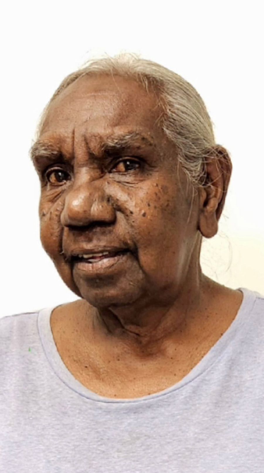 Elderly Aboriginal woman in a light blue shirt, smiling. White background.