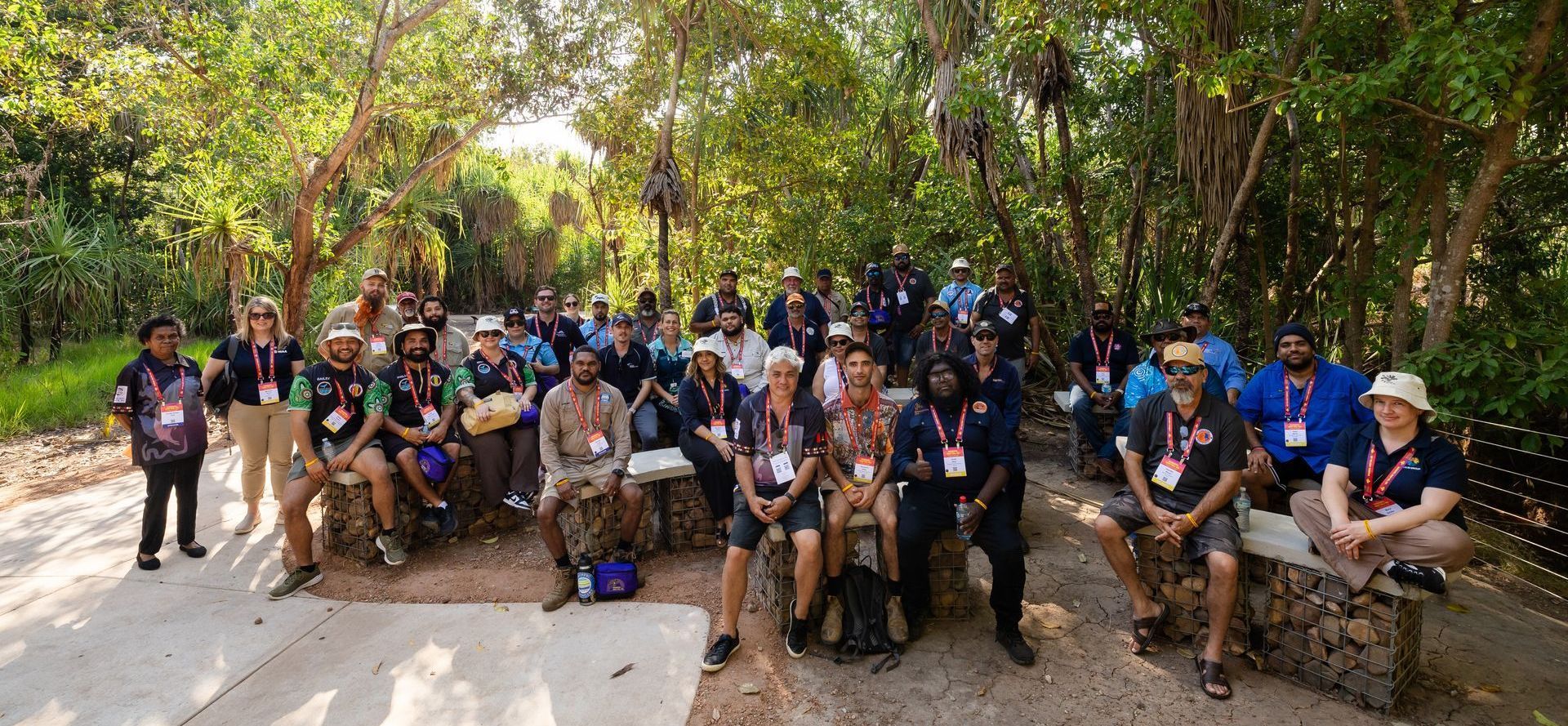 Group of people in a lush, green outdoor setting, some sitting, some standing, all wearing lanyards, possibly on a tour.