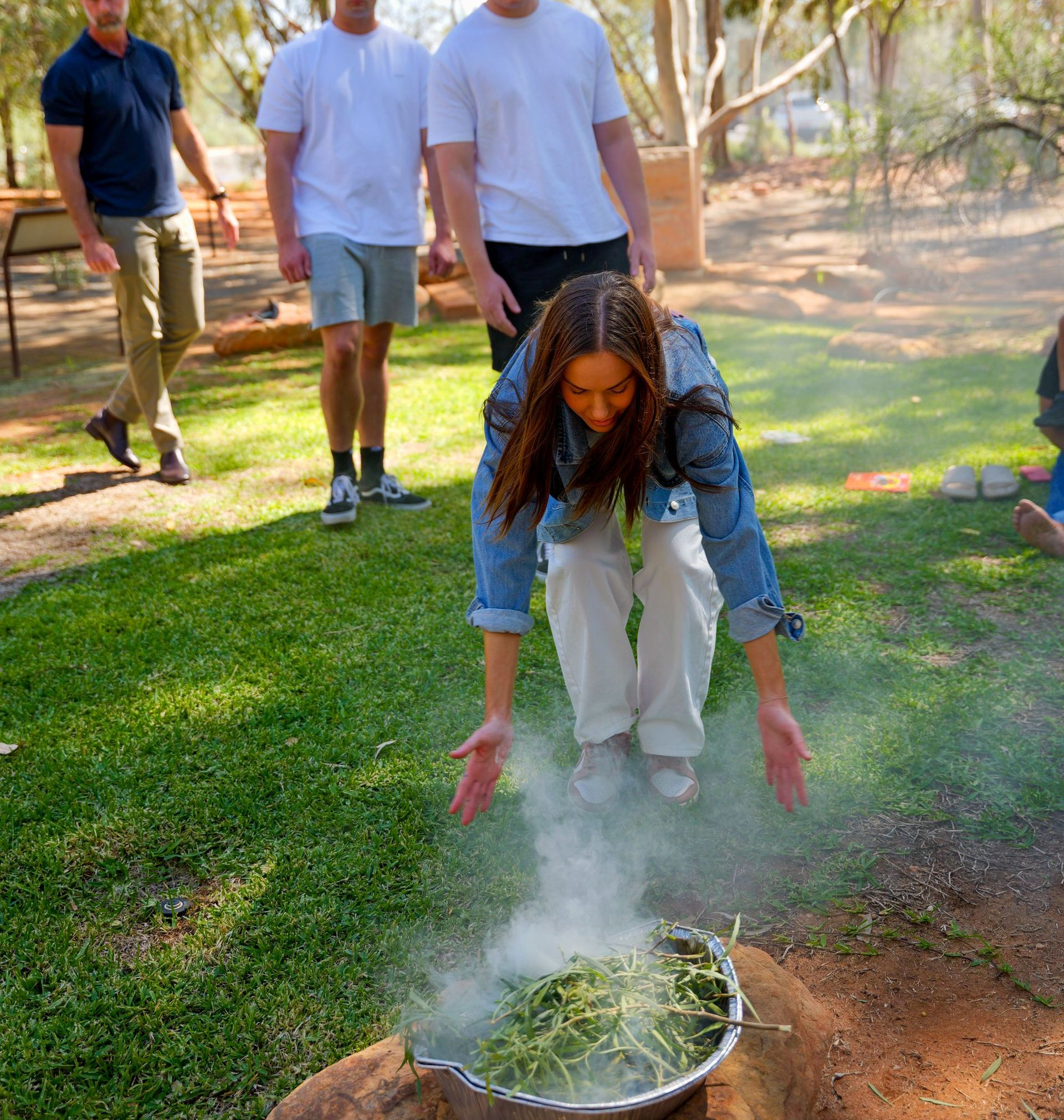 Woman bends over a smoking bowl outdoors while three people watch in a sunny garden