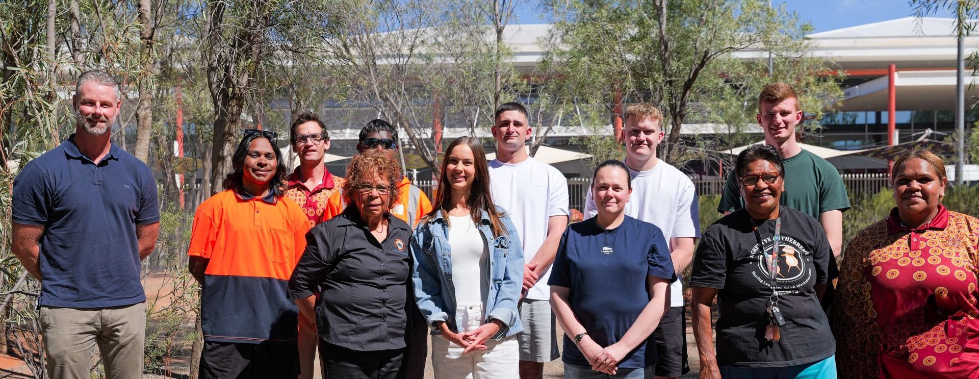 Group of people standing outdoors in front of buildings and trees on a sunny day.