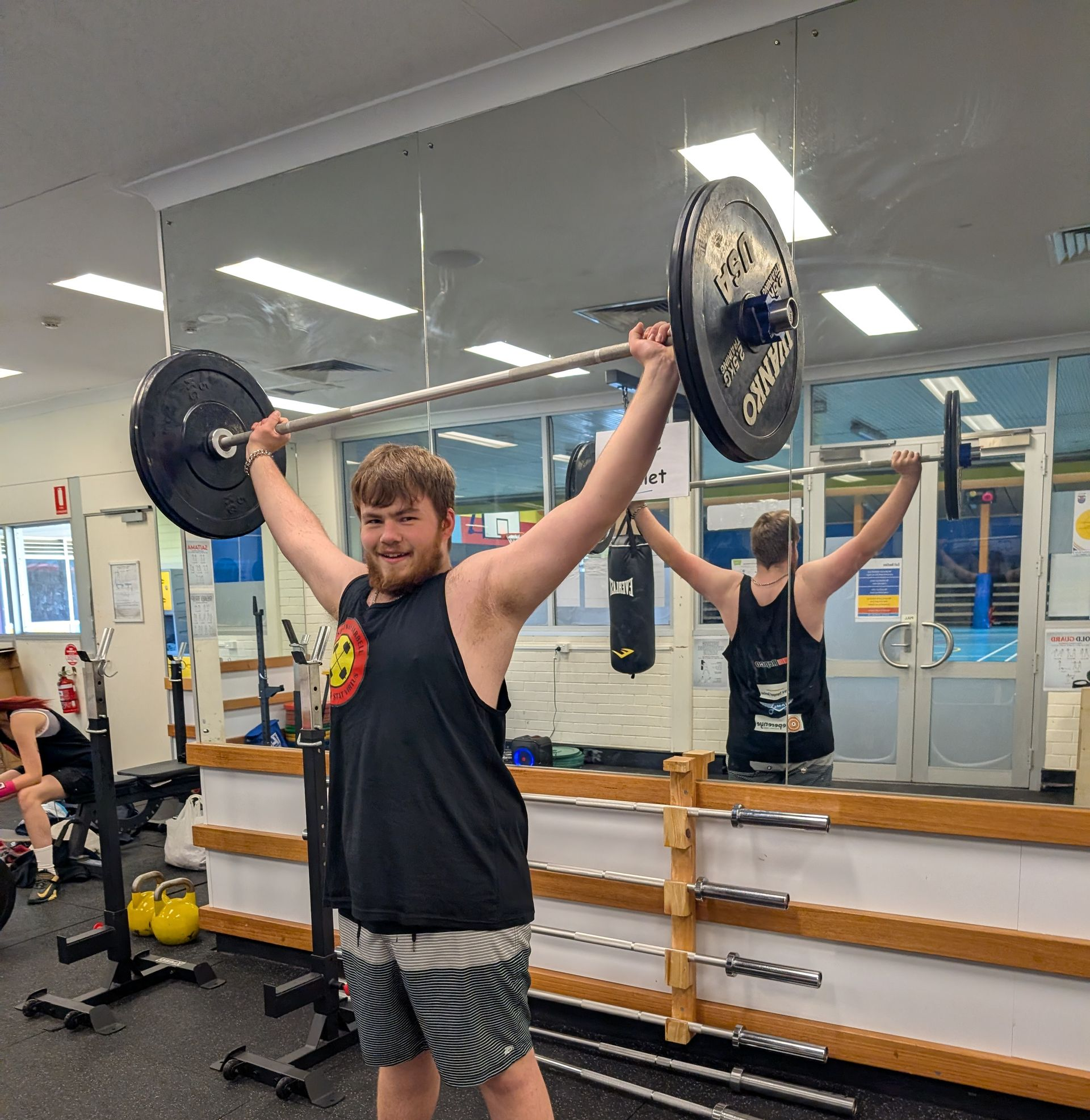 Man lifting barbell overhead in a gym, arms extended, black shirt, shorts, mirror in background.