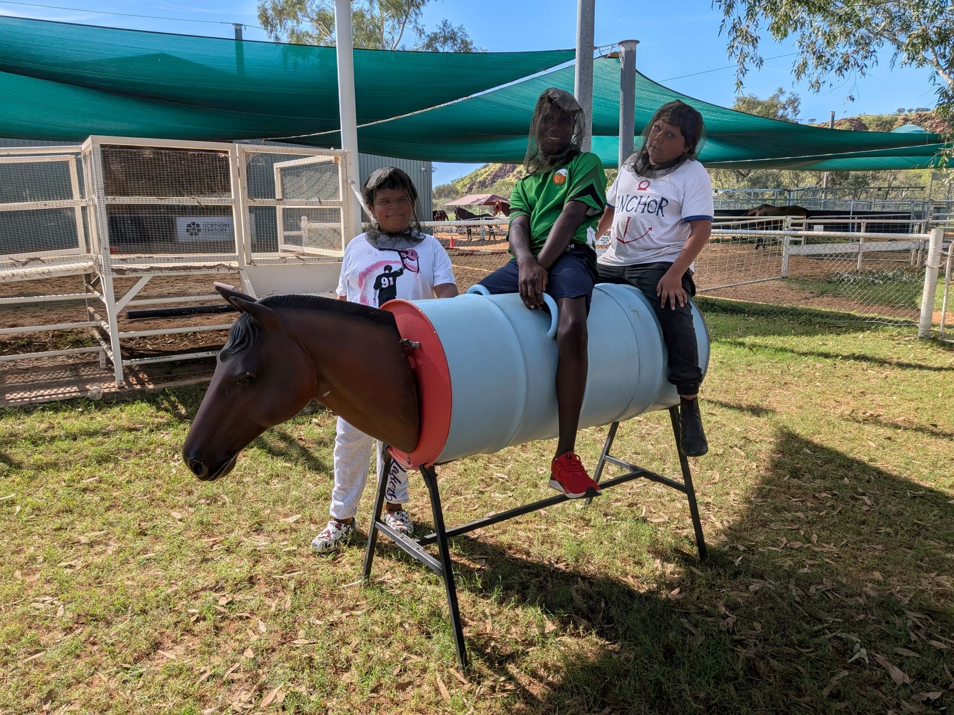 Three children sit on a painted horse ride in a sunny fenced outdoor play area.