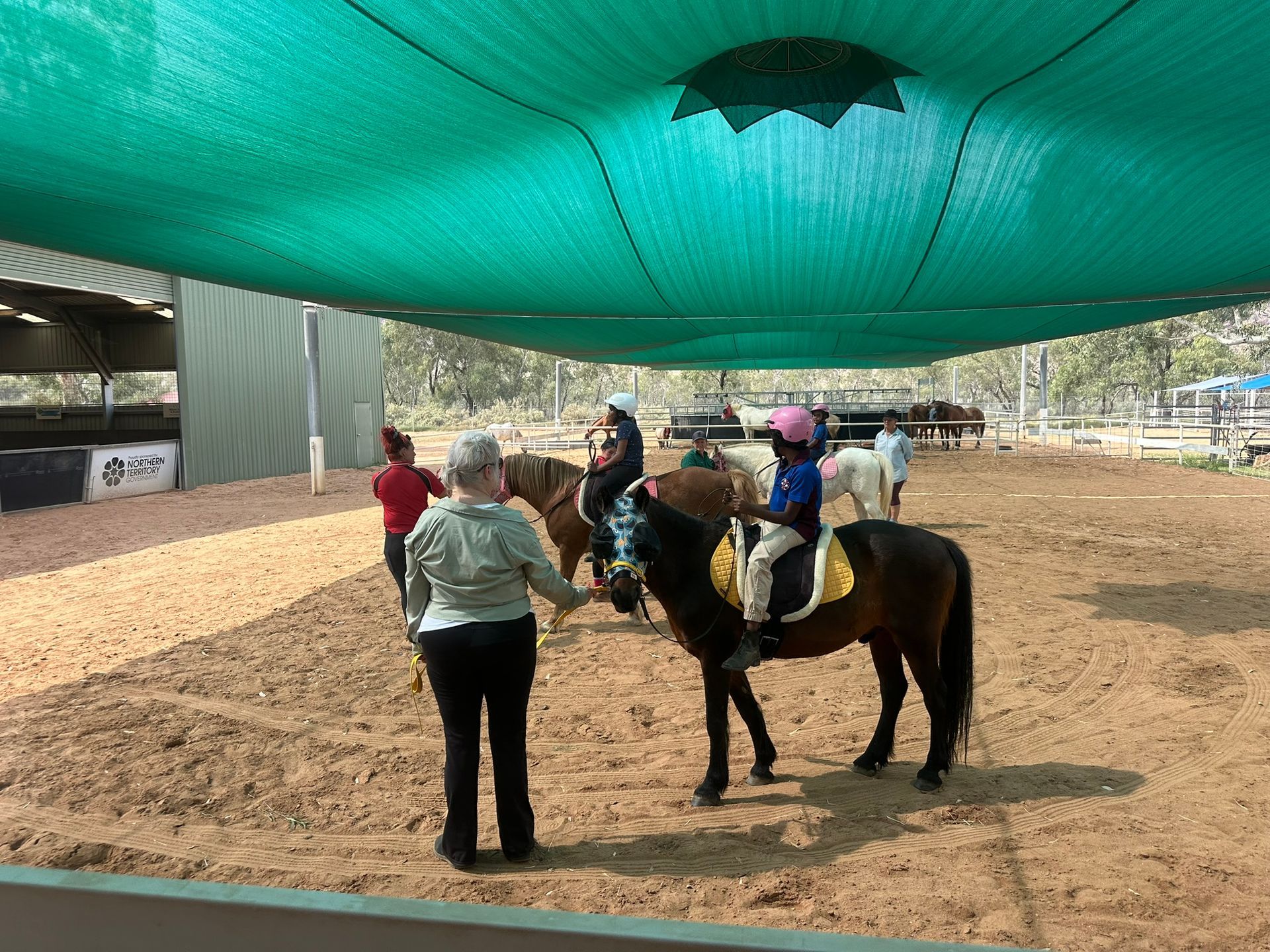 People riding horses in an indoor arena under a green canopy, with an instructor nearby