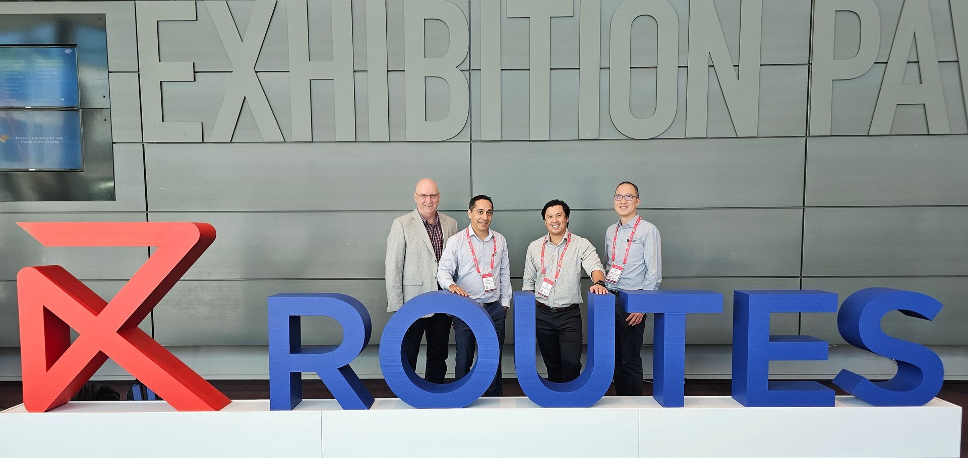 Four men standing behind large blue letters spelling ROUTES, with a red X in front. Grey background reads EXHIBITION PARK.