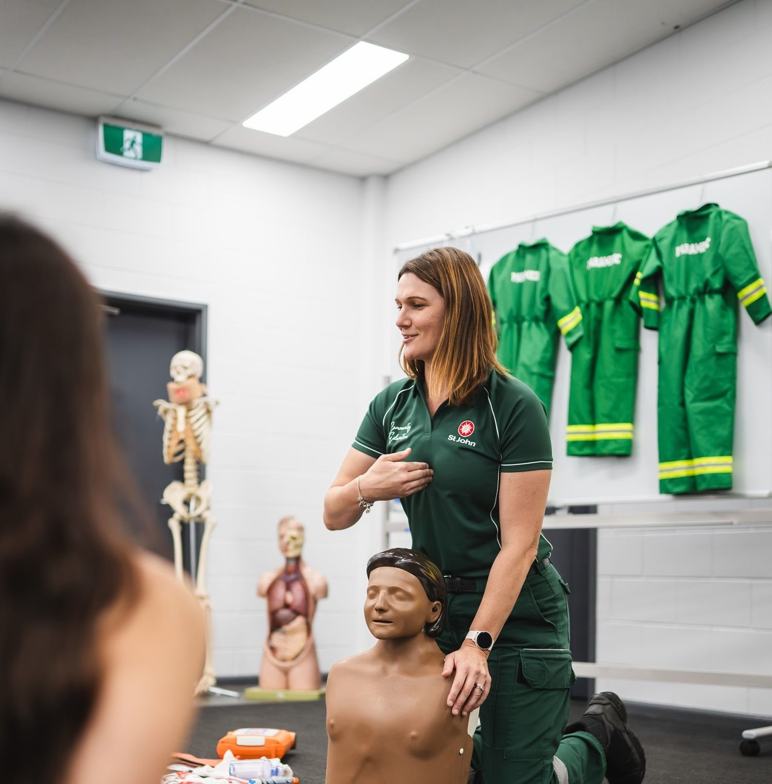 Woman in green uniform demonstrating CPR on a mannequin in a classroom setting.