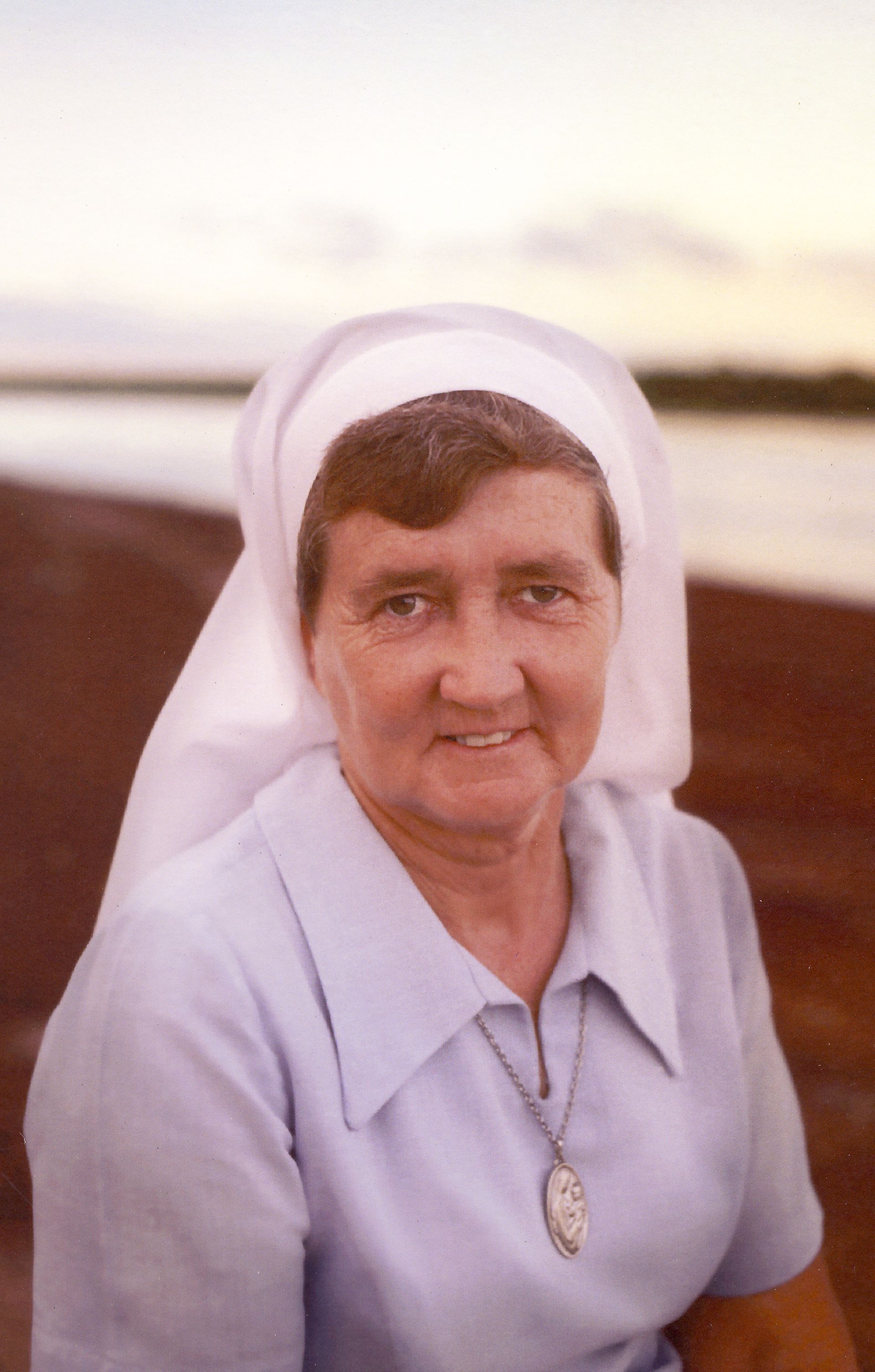 Nun in white habit and blue shirt smiles at the camera, in a blurry outdoor setting.