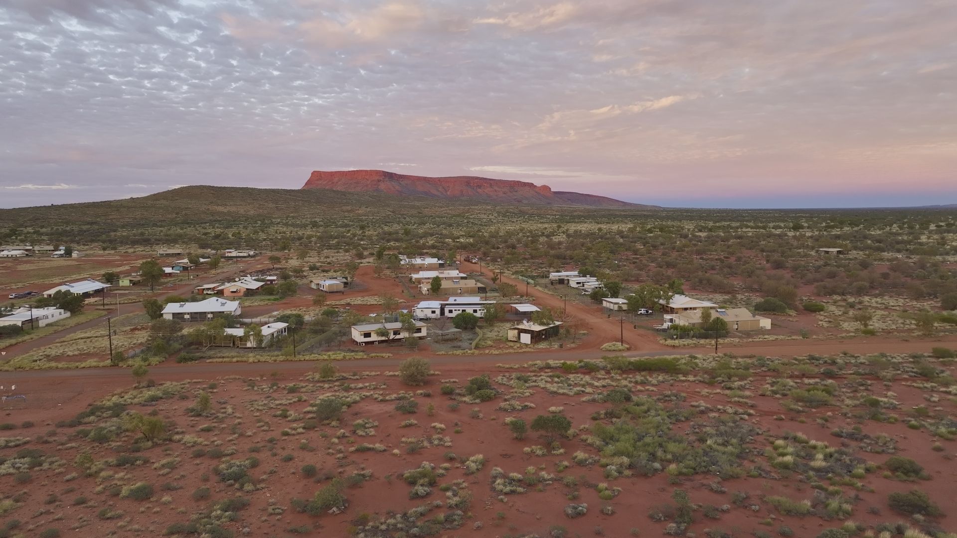 Outback town with red earth, scattered buildings, and a mesa under a colorful, dusky sky.