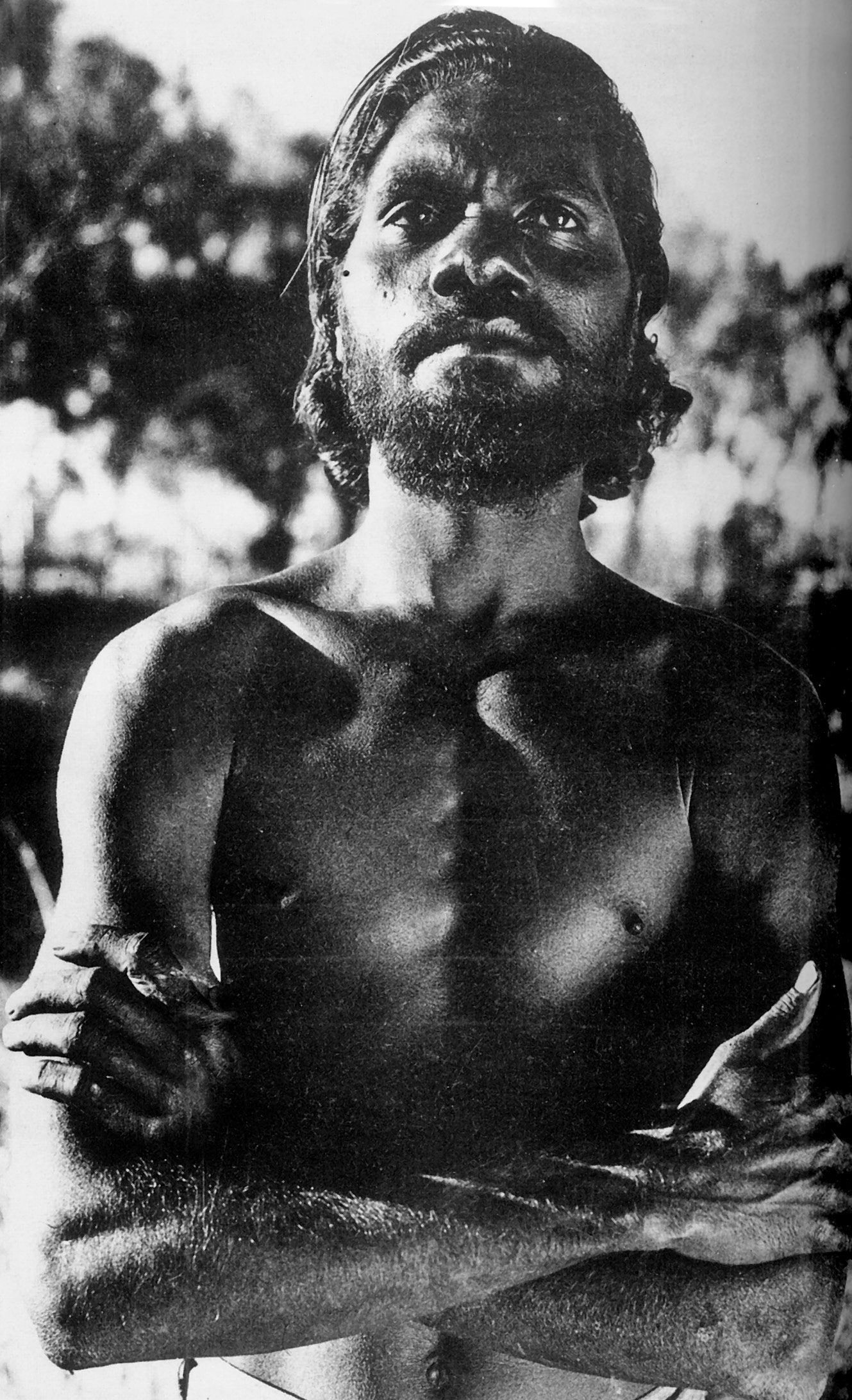 Aboriginal man standing with arms crossed, gazing forward. Outdoors, shirtless, with a beard.