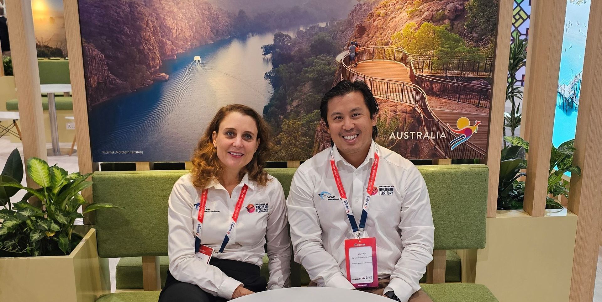 Two people sit smiling in front of a scenic photo, wearing white shirts and lanyards.
