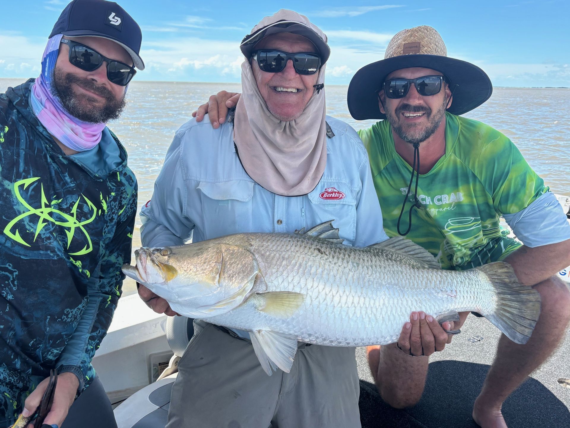 Three people smiling on a boat hold a large silver fish over the water.