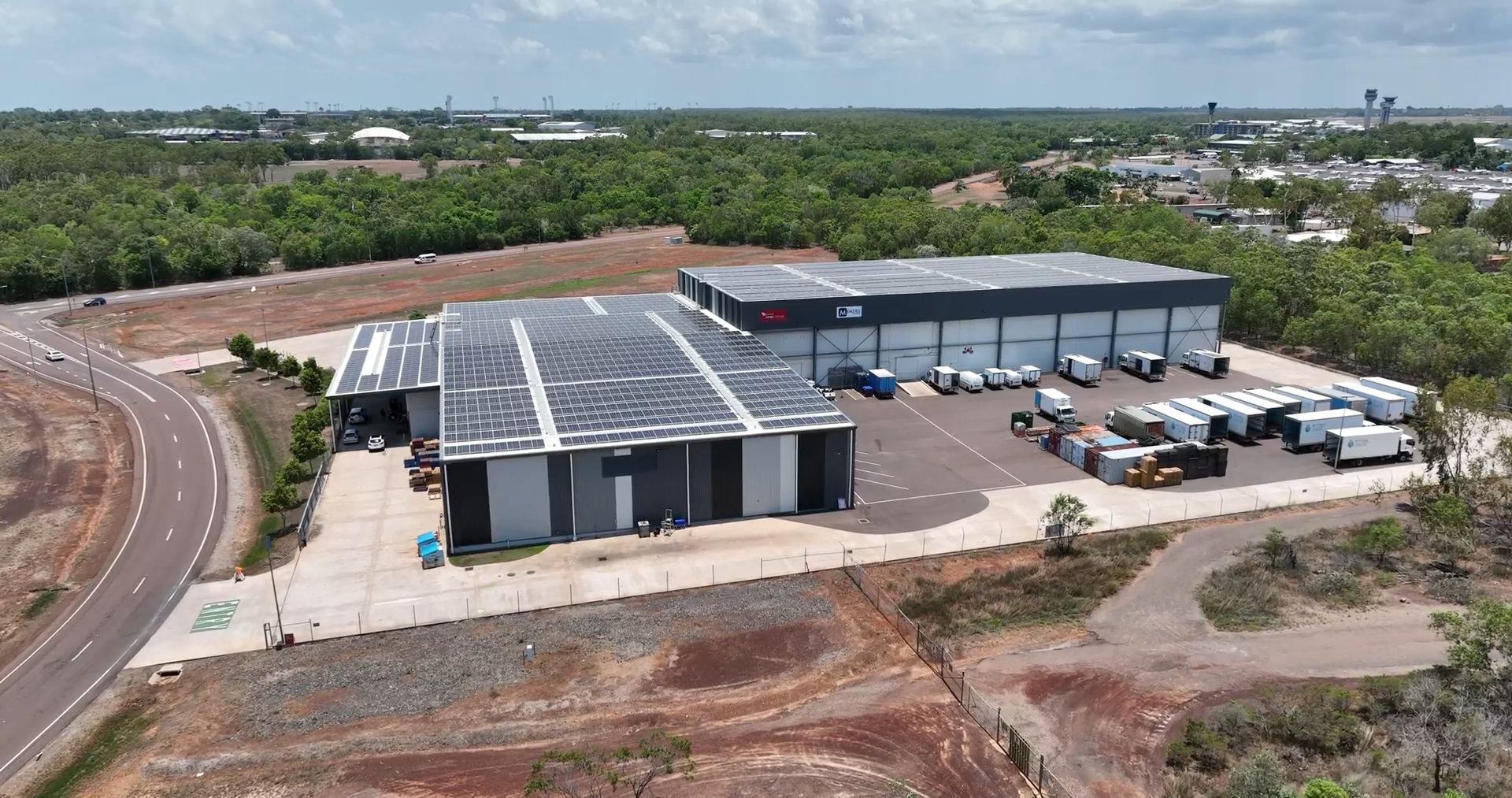 Aerial view of a warehouse complex with trucks parked outside. Red dirt and trees surround the building.