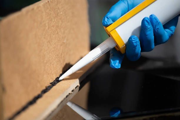A person wearing blue gloves applies black sealant from a caulk gun to a cardboard structure.