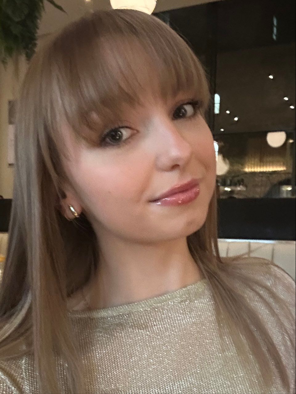 Blonde woman with bangs, smiling at camera, wearing gold top, indoors.