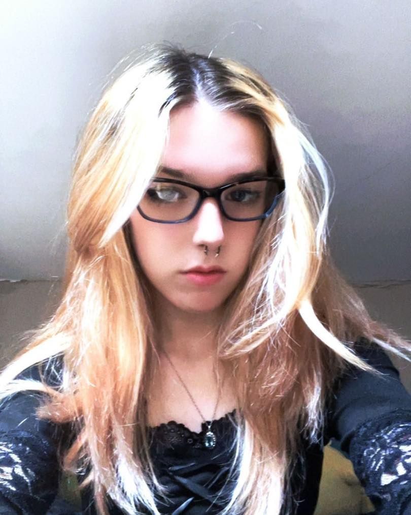 Person with long, blonde and brown hair, wearing black glasses and a nose ring, with a black top.