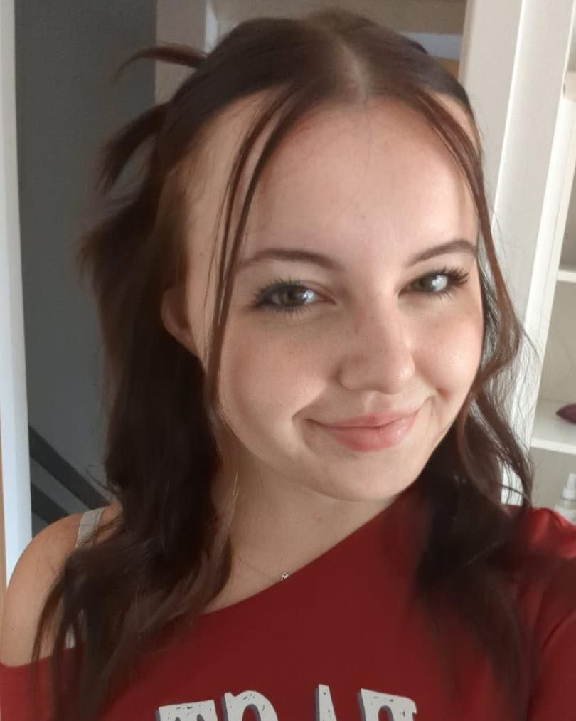 Woman with brown hair smiling, wearing a red shirt, looking at the camera. White wall in background.