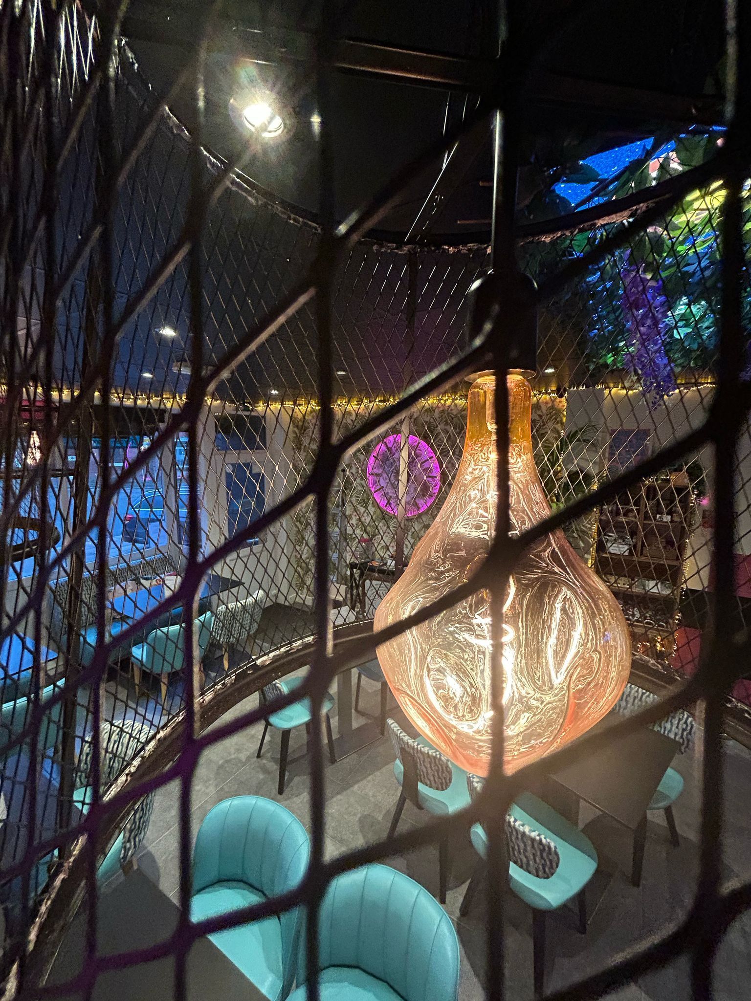 View of a decorated venue through a dark metal gate. Teal chairs, tables, and a large, glowing light fixture are visible.