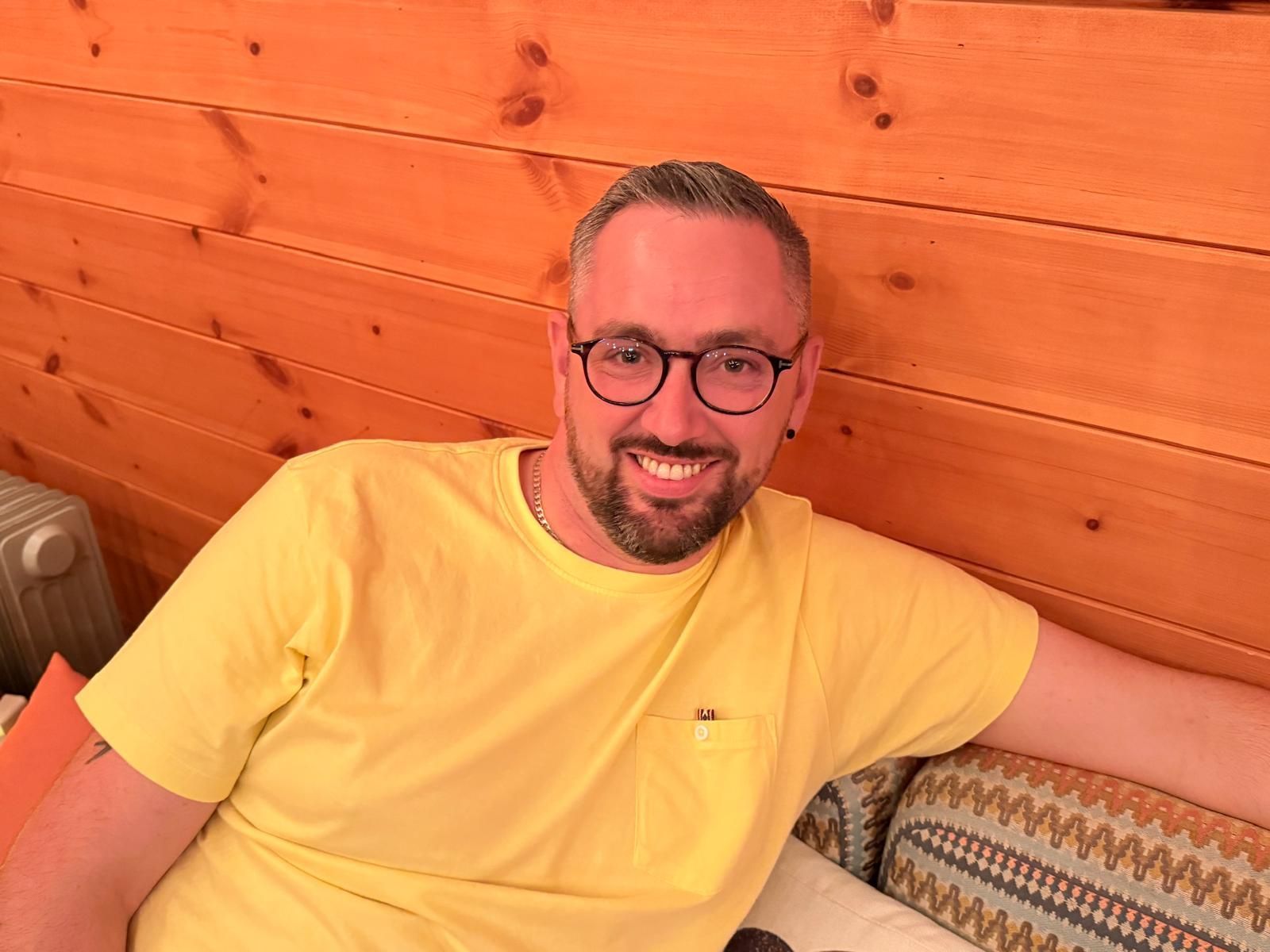 Man with glasses and yellow shirt smiles, seated on a couch in front of wood paneling.