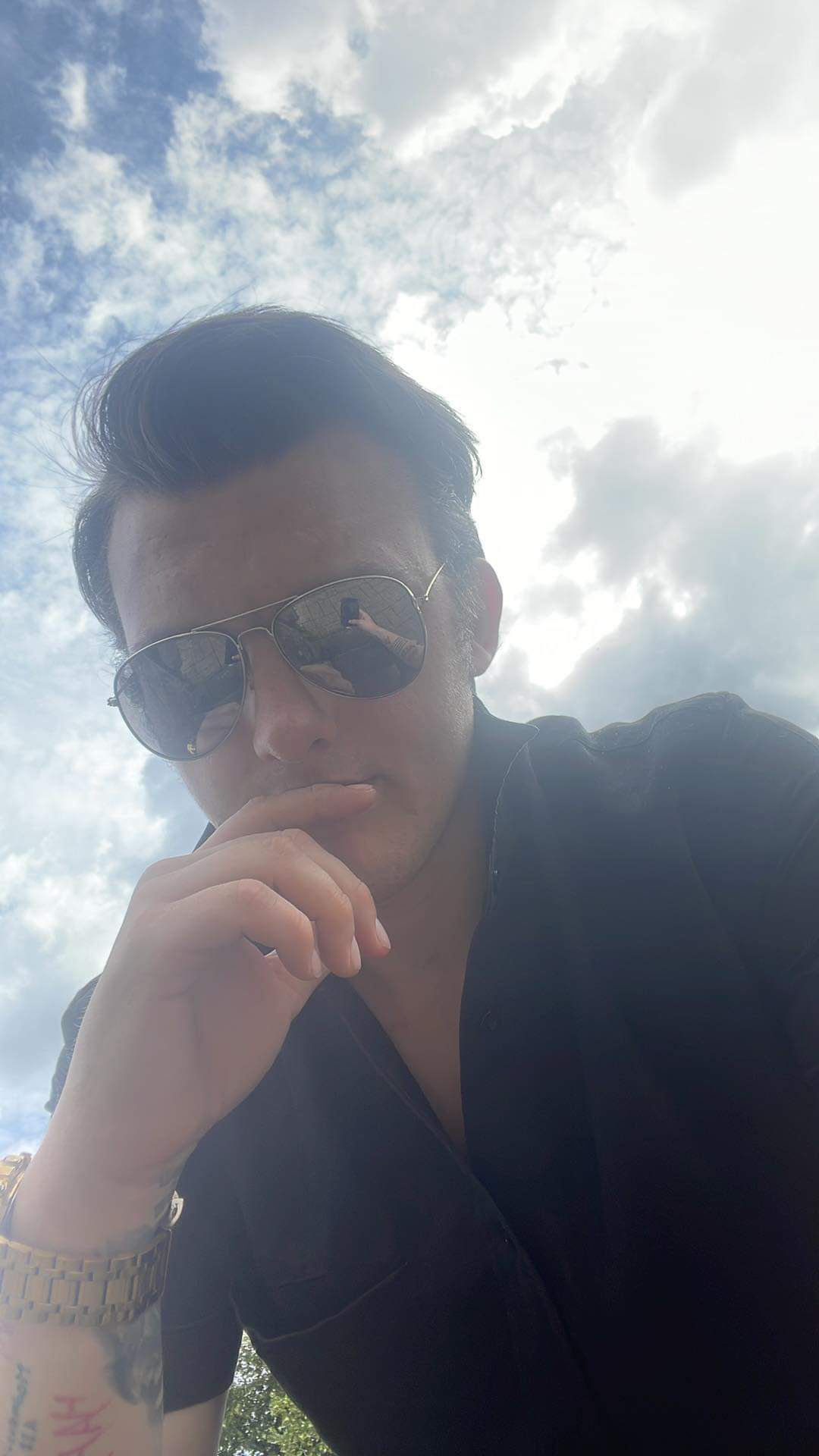 Man in sunglasses, black shirt, hand on chin, against cloudy sky.