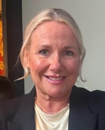 Blond woman wearing a black blazer smiles at the camera indoors.