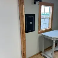 A wall-mounted black safe sits next to a window with wooden trim in a room with a light gray table.