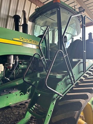 Green John Deere tractor cab with metal safety railings and stairs, parked inside a barn.