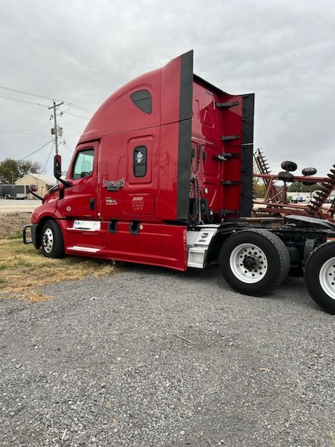 A red Freightliner semi-truck parked on a gravel lot under a cloudy sky.