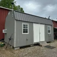 A grey, barn-style shed with a metal roof, white double doors, two windows, and an air conditioning unit on the side.