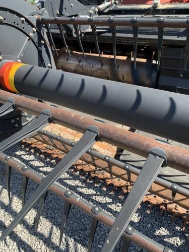 A close-up of a combine harvester's reel, showing black metal tines and a black tube with yellow and orange bands.