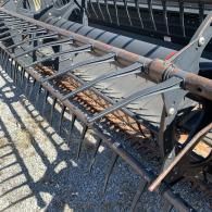 Close-up view of the metal reel and pick-up tines of a combine harvester header, angled on a gravel surface.