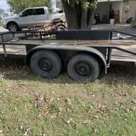 A black flatbed utility trailer with two axles sits on grass near a parked white pickup truck.