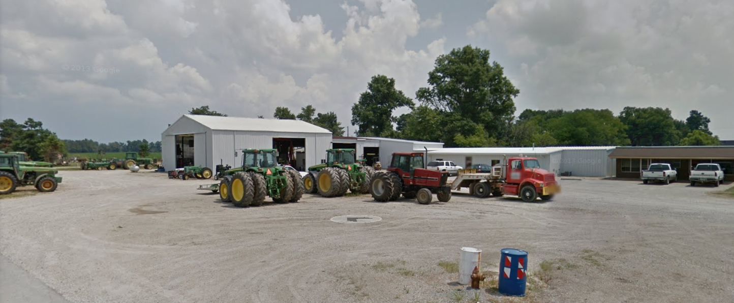 A collection of green, red, and yellow tractors parked on a gravel lot in front of white industrial warehouse buildings.