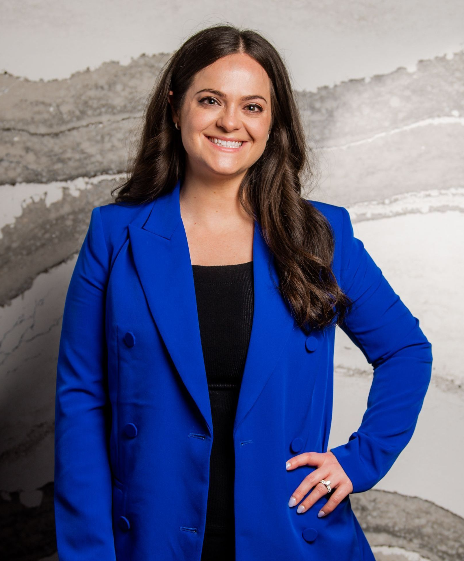Woman in a blue blazer smiles, hand on hip, against a marble wall.