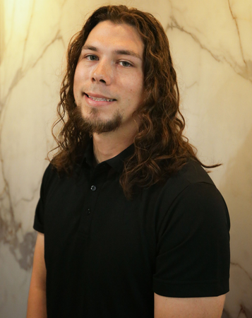 Smiling person with long hair wearing a black shirt, standing against a beige marbled wall.