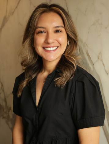Smiling person in a black blouse standing against a beige marble wall.
