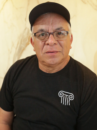 Man wearing a black cap and black T-shirt with a white logo, standing against a light marble wall.