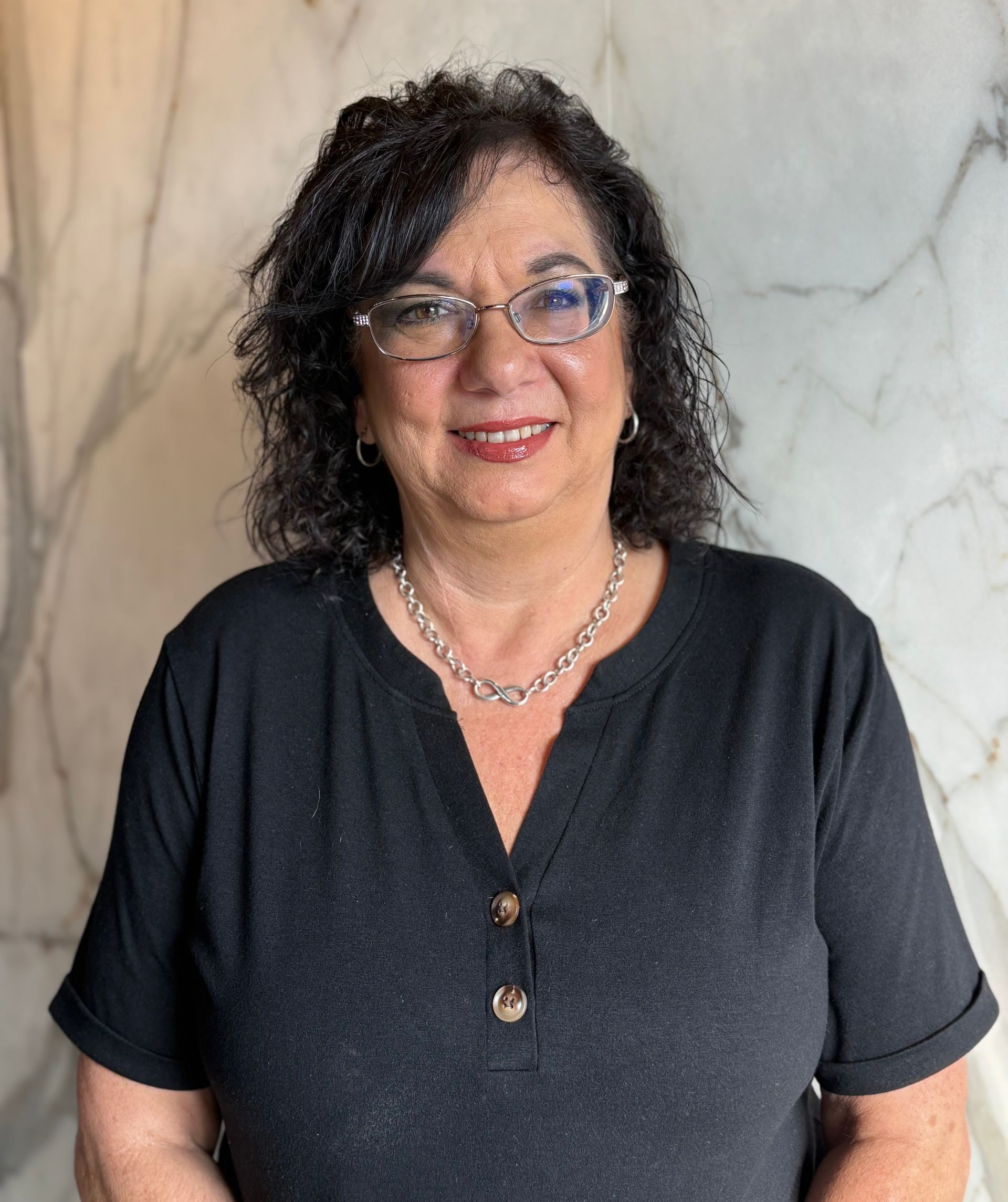 Woman wearing glasses smiles, wearing a black shirt, and a beaded necklace, in front of a white marble wall.